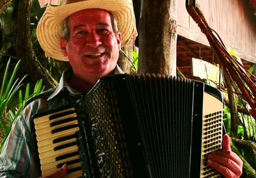 Foto Dino Rocha Tocando acordeon sorrindo, ao fundo uma casa rural.